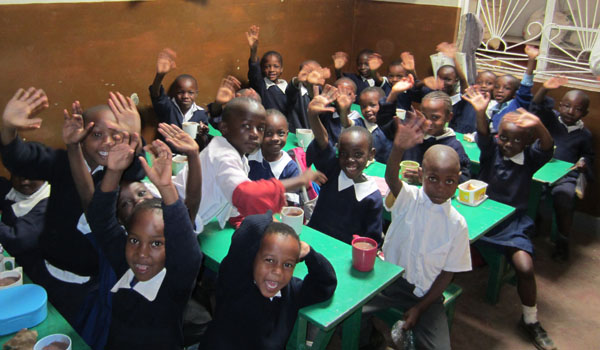 Children seated together at green classroom tables and waving.